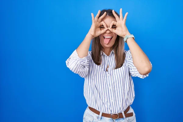 西班牙裔年轻女子站在蓝色的背景上做着像双筒望远镜一样的手势 伸出舌头 眼睛透过手指看着 疯狂的表达 — 图库照片