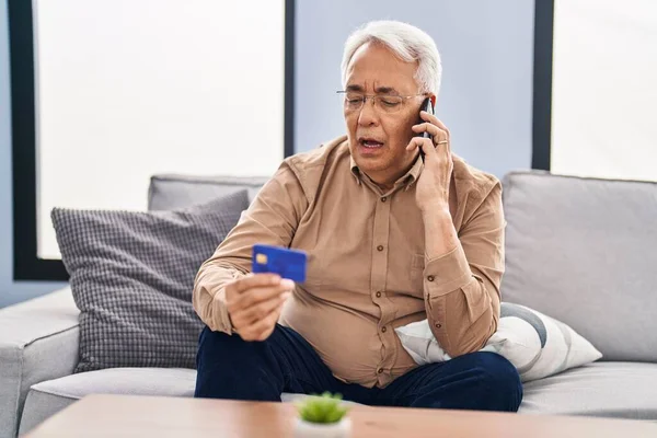 Senior man talking on the smartphone holding credit card sitting on sofa at home