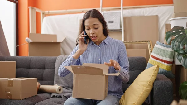 Young beautiful hispanic woman unpacking cardboard box speaking on the phone complaining at new home