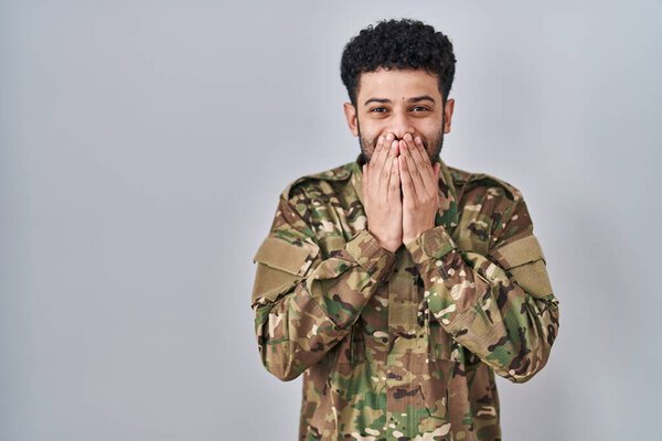 Arab man wearing camouflage army uniform laughing and embarrassed giggle covering mouth with hands, gossip and scandal concept 