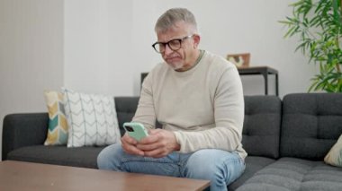 Orta yaşlı, kır saçlı bir adam evde stresli bir ifadeyle akıllı telefon kullanıyor.