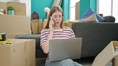 Genç sarışın kadın dizüstü bilgisayar kullanıyor. Yeni evinde akıllı telefondan konuşuyor.