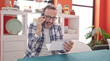 Orta yaşlı bir adam yemek odasında akıllı telefondan konuşan öfkeli bir evrak okuyor.