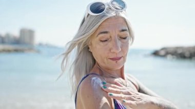 Orta yaşlı, kır saçlı, bikini giyen turist plajda güneş kremi sürüyor.