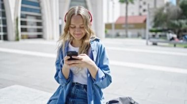 Üniversitede akıllı telefon ve kulaklık kullanan genç sarışın kız öğrenci.