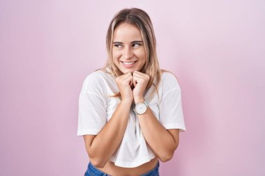 Pembe arka planda duran genç sarışın kadın. Gergin ve heyecanlı, elleri çenesinde yan tarafa bakıyor. 