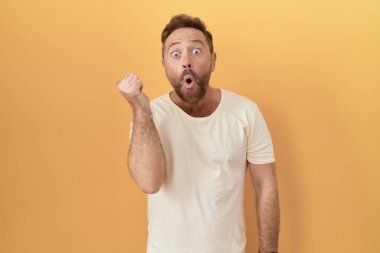 Sarı arka planda dikilen sakallı orta yaşlı bir adam elleri yana doğru işaret ederek şaşırdı, ağzı açık hayret içinde kaldı.. 