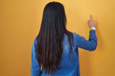 Sarı arka planda duran genç Asyalı kadın parmak ile öne doğru poz veriyor. 