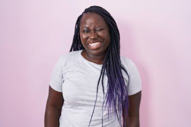 Genç Afrikalı bir kadın pembe arka planda duruyor. Seksi bir ifadeyle kameraya bakıyor. Neşeli ve mutlu bir yüzü var.. 