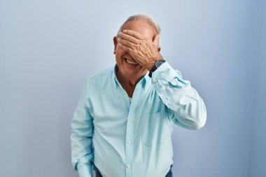 Mavi arka planda gri saçlı, gülümseyen ve gülümseyen, yüzünde şaşkınlıktan gözleri kapayan son sınıf öğrencisi. kör kavram. 