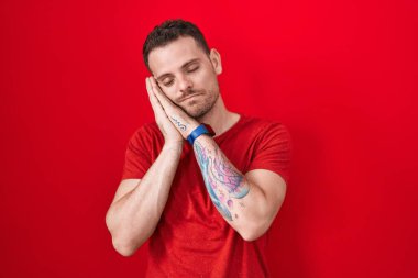 Young hispanic man standing over red background sleeping tired dreaming and posing with hands together while smiling with closed eyes. 