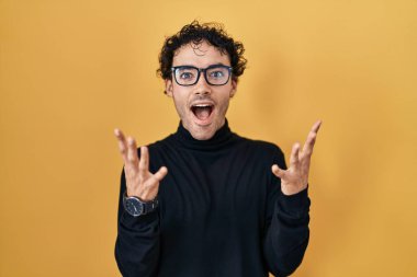 Hispanic man standing over yellow background celebrating crazy and amazed for success with arms raised and open eyes screaming excited. winner concept 