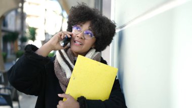 Genç Afrikalı Amerikalı kadın gülümsüyor. Elinde klasörle caddede telefonla konuşuyor.