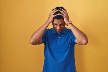 Young hispanic man standing over yellow background suffering from headache desperate and stressed because pain and migraine. hands on head. 