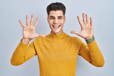 Young hispanic man standing over blue background showing and pointing up with fingers number ten while smiling confident and happy. 