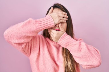 Young hispanic woman standing over pink background covering eyes and mouth with hands, surprised and shocked. hiding emotion 