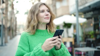 Young blonde woman smiling confident using smartphone at coffee shop terrace