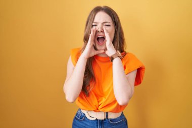 Caucasian woman standing over yellow background shouting angry out loud with hands over mouth  clipart