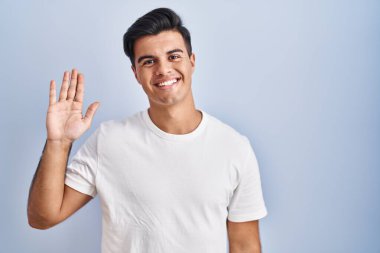 Hispanic man standing over blue background waiving saying hello happy and smiling, friendly welcome gesture 