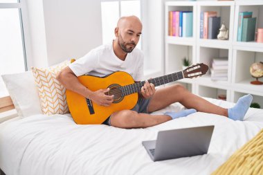 Genç kel bir adamın online klasik gitar dersi var. Yatak odasında oturuyor.