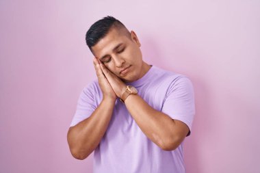 Young hispanic man standing over pink background sleeping tired dreaming and posing with hands together while smiling with closed eyes. 