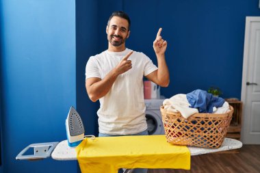 Sakallı, evde ütü yapan, gülümseyen ve kameraya bakan, iki eli ve parmağı yanda olan genç İspanyol bir adam.. 