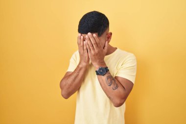 Sarı arka planda dikilen genç İspanyol adam. Ağlarken suratını ellerle kapatıyor. depresyon kavramı. 