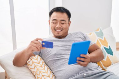 Young chinese man using touchpad and credit card sitting on bed at bedroom