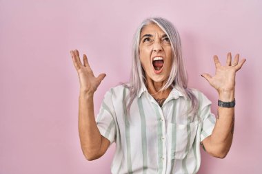 Middle age woman with grey hair standing over pink background crazy and mad shouting and yelling with aggressive expression and arms raised. frustration concept. 