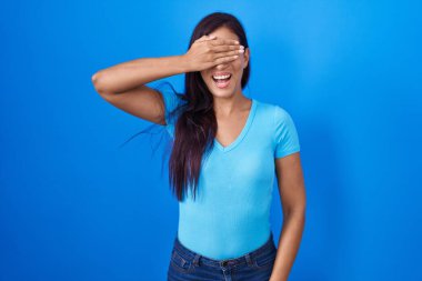 Mavi arka planda dikilen genç İspanyol kadın. Gülümsüyor ve gülümsüyor. Gözlerini şaşkınlıkla kapatıyor. kör kavram. 