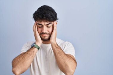 Beyaz arka planda duran sakallı İspanyol bir adam, eli başının üstünde, stres yüzünden başı ağrıyor. migren ağrısı. 