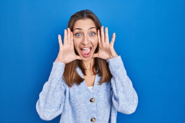 Young woman standing over blue background smiling cheerful playing peek a boo with hands showing face. surprised and exited 