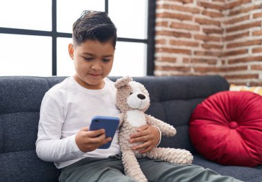 Adorable hispanic boy using smartphone sitting on sofa at home