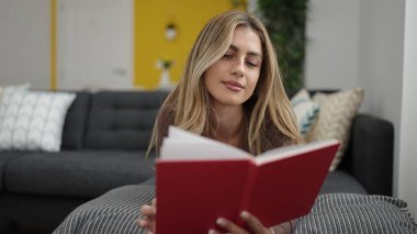 Young blonde woman reading book lying on sofa at home