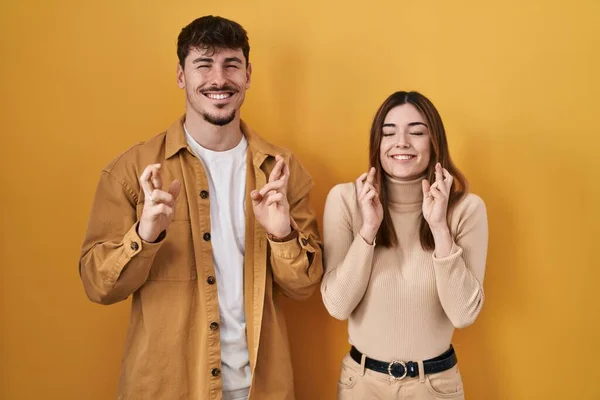 若いヒスパニック系のカップルが黄色の背景ジェスチャー指の上に立って希望と目を閉じて笑顔を渡った 幸運と迷信の概念は — ストック写真