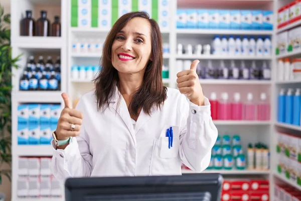 薬局で働く中世のブルネットの女性薬局の成功サイン手で積極的なジェスチャーをし 親指を上げて笑顔と幸せ 陽気な表情と勝者のジェスチャー — ストック写真