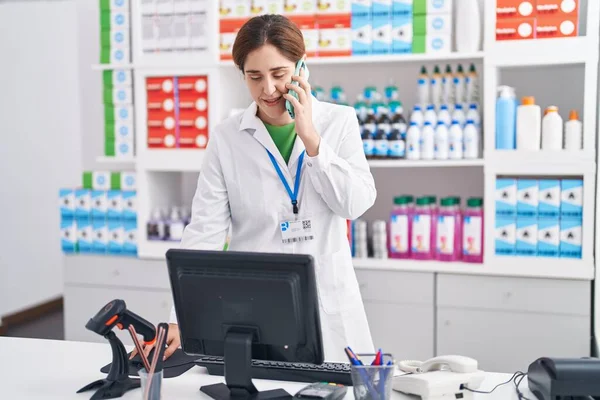 stock image Young woman pharmacist talking on smartphone using computer at pharmacy