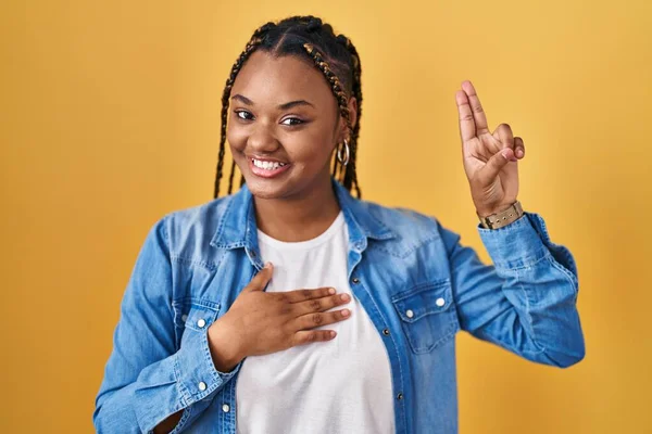 African american woman with braids standing over yellow background smiling swearing with hand on chest and fingers up, making a loyalty promise oath