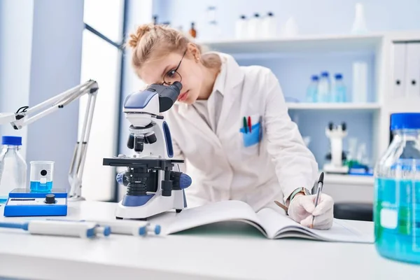 Young Blonde Woman Scientist Using Microscope Writing Report Laboratory — Foto de Stock