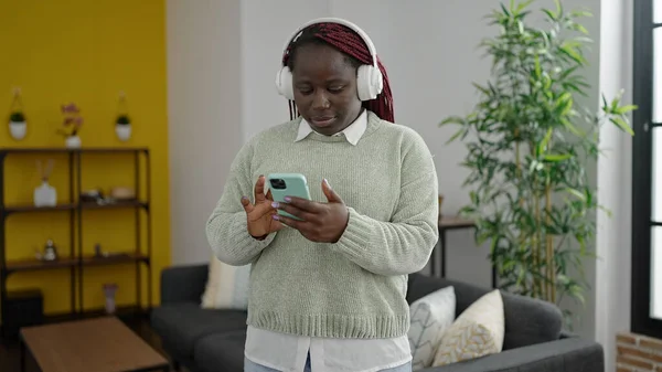 在家里戴耳机用智能手机剪辫子的非洲妇女 — 图库照片