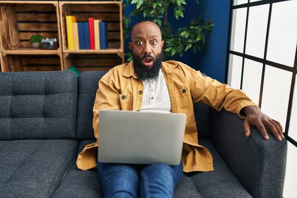 アフリカ系アメリカ人男性使用してノートパソコンを自宅でソファの上に座って怖がって驚きのために口を開けて 不信の顔 — ストック写真