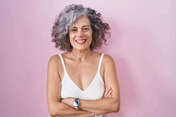 Middle age woman with grey hair standing over pink background happy face smiling with crossed arms looking at the camera. positive person.