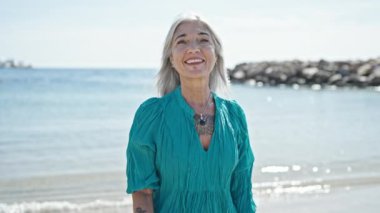 Orta yaşlı, gri saçlı, gülümseyen, kendine güvenen bir kadın kumsalda el kol hareketi yapıyor.