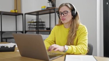 Ofiste çalışan genç sarışın iş kadını dizüstü bilgisayar ve kulaklık kullanıyor.
