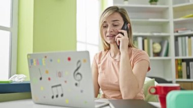 Kütüphanede dizüstü bilgisayar kullanan genç sarışın kız öğrenci akıllı telefondan konuşuyor.