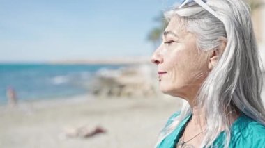 Orta yaşlı, kır saçlı bir kadın plajda rahat bir ifadeyle dikiliyor.