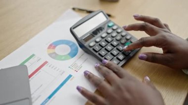 Saçları örgülü Afrikalı bir kadın ofiste dizüstü bilgisayar ve hesap makinesi kullanıyor.