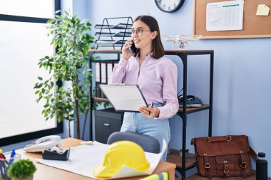 Young hispanic woman architect talking on the smartphone reading document at office