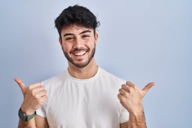 Hispanic man with beard standing over white background success sign doing positive gesture with hand, thumbs up smiling and happy. cheerful expression and winner gesture. 