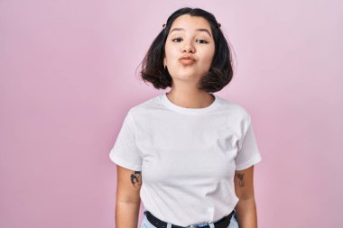 Latin kökenli genç bir kadın pembe arka plan üzerine gündelik beyaz t-shirt giyip kameraya bakıp havada öpüşerek güzel ve seksi görünmeye çalışıyor. aşk ifadesi. 